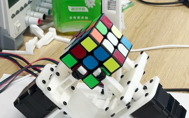 The dual-arm robot holding an unsolved Rubik's cube corner-down between its two white printed grippers